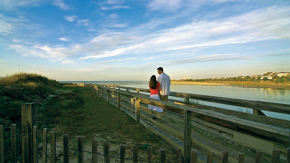 A couple walks on a wooden boardwalk leading to the beach.