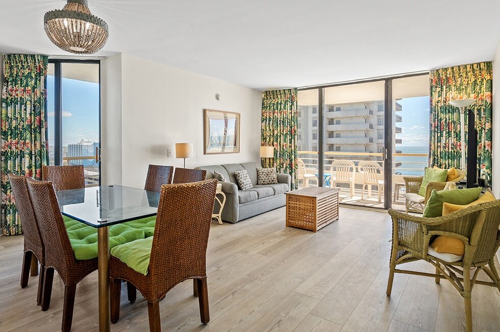 A wide view of a dining area with a glass table and wicker chairs, adjacent to a living room with a sofa and balcony.