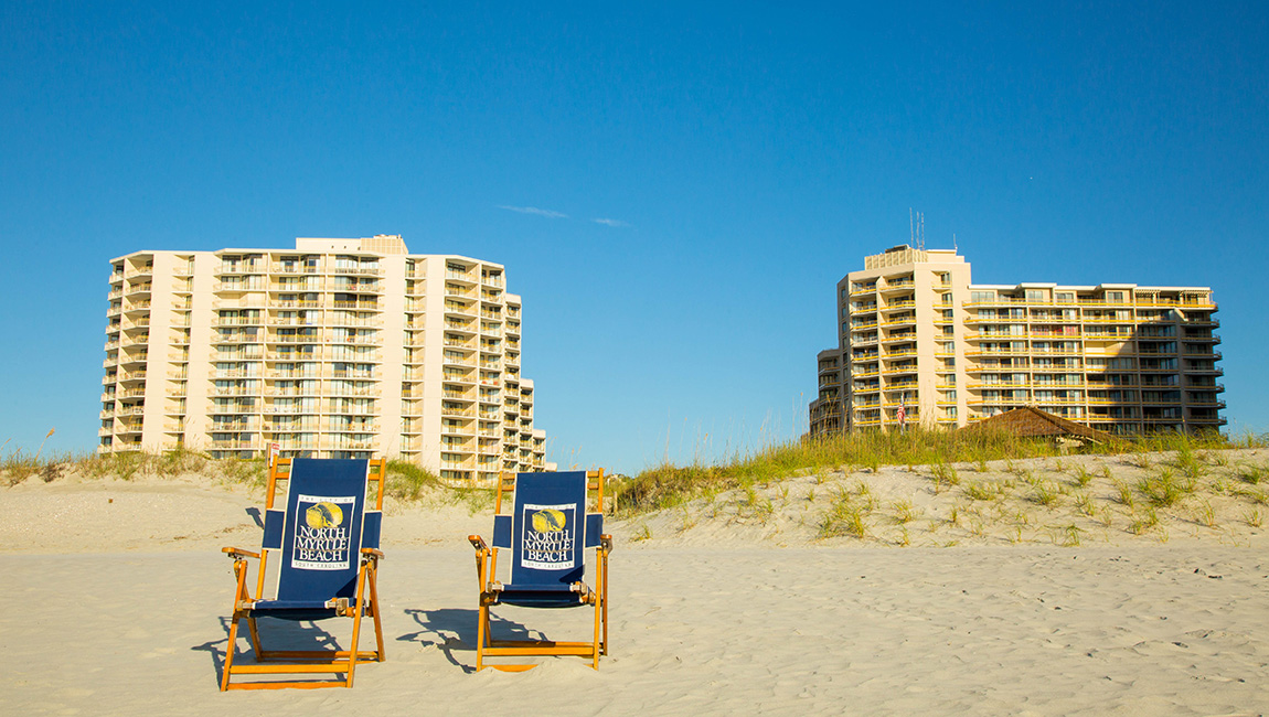 Two beach chairs on a sandy beach with two large buildings in the background.
