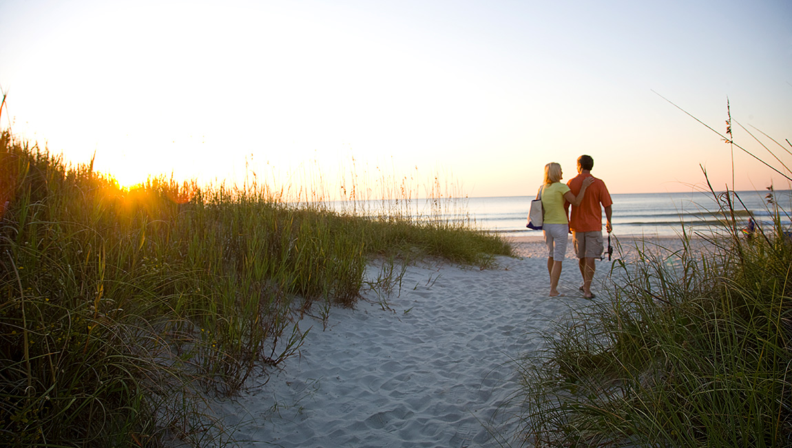 A couple walks along a beach path at sunset.
