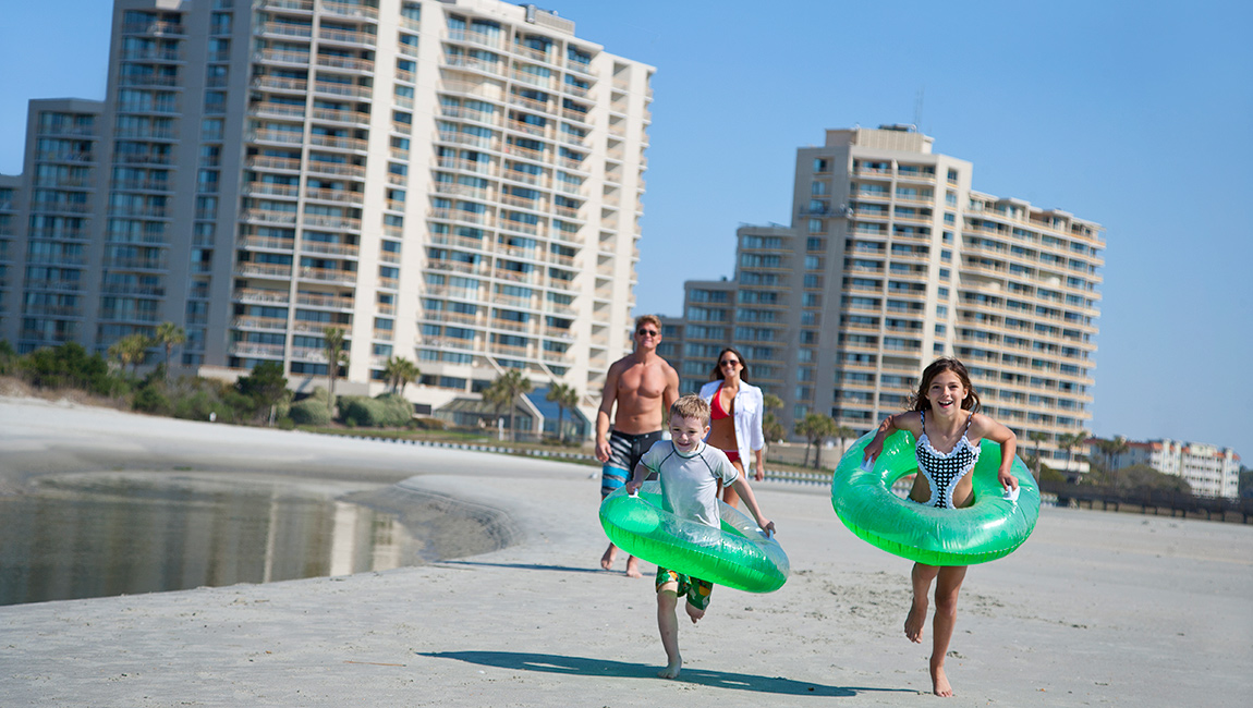 A family is running on the beach with inflatable floats in front of a large hotel.