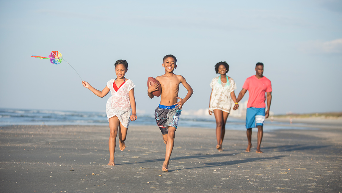 A family of four is running on the beach, with a kite and football.