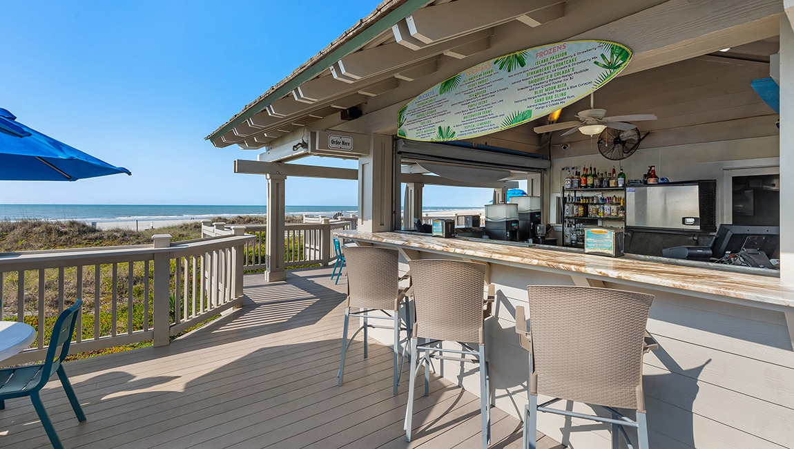 A beachside bar with a menu board, bar stools, and a deck overlooking the ocean.