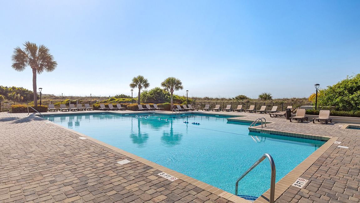 A large rectangular pool with a brick paver deck, surrounded by lounge chairs and palm trees under a clear blue sky.