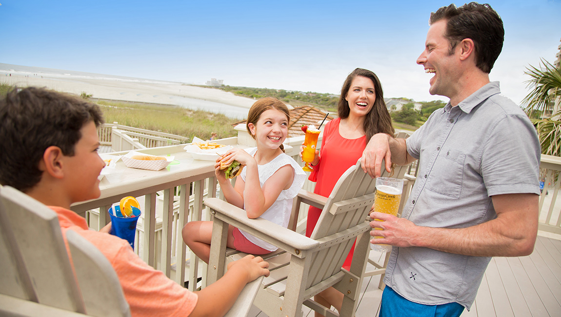 A family of four enjoys a meal on a balcony overlooking the beach.