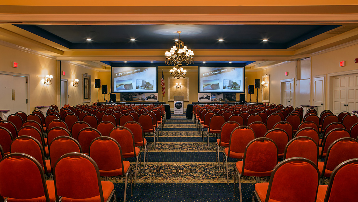 A large meeting room with rows of red chairs facing a projector screen at the front of the room.