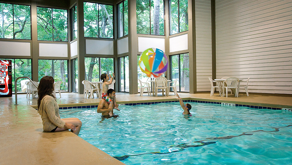 A family enjoys playing with a large beach ball in an indoor pool surrounded by windows showing trees outside.