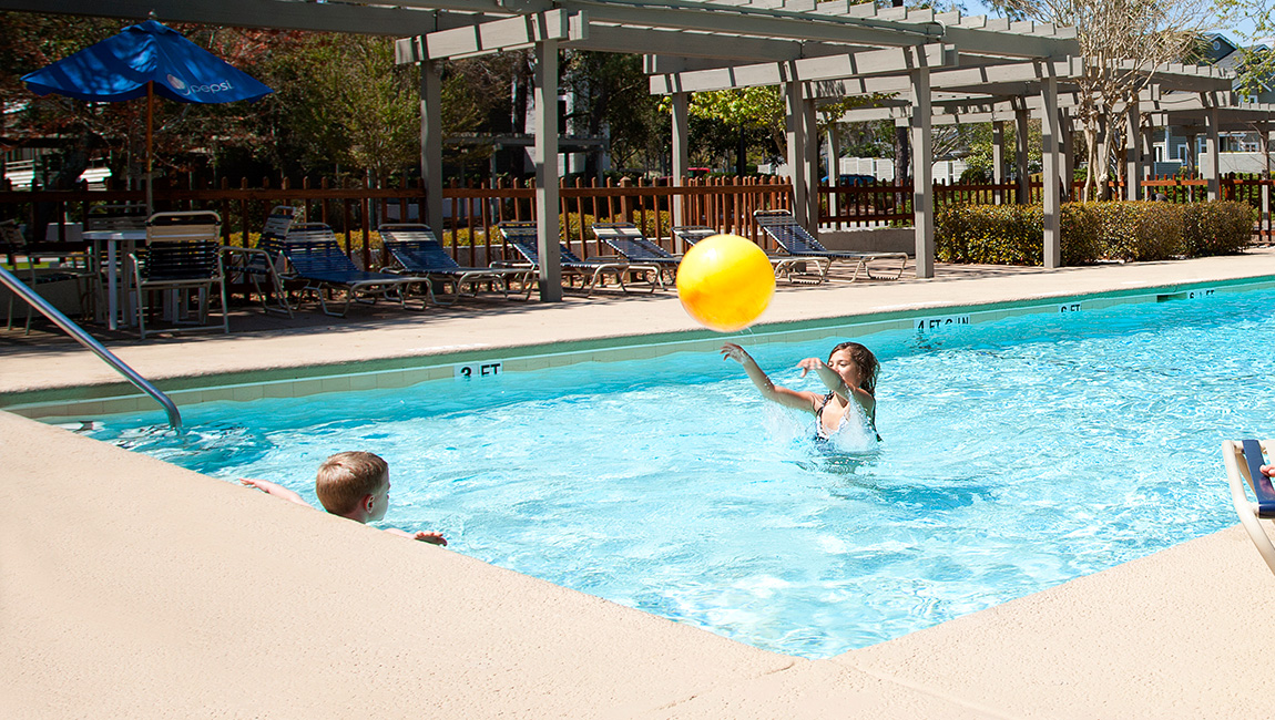 A woman and a child playing with a yellow ball in a swimming pool with a pergola and lounge chairs in the background.