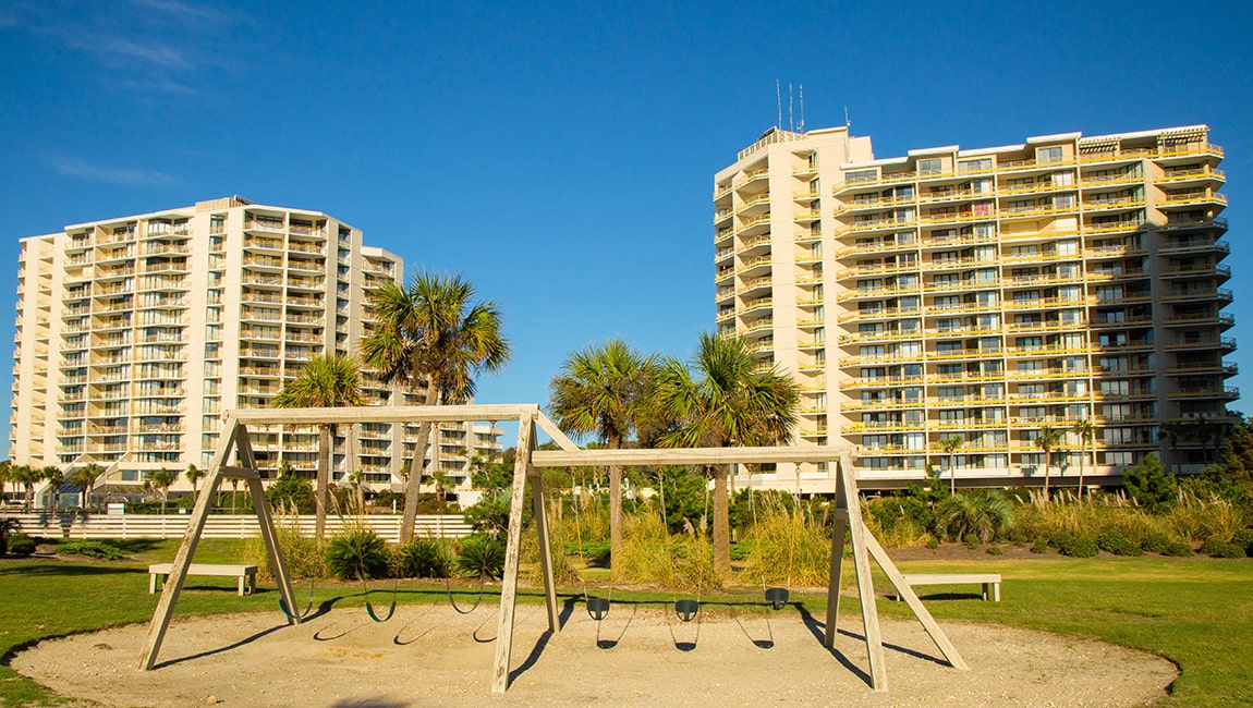 A wide-angle view of a beachfront resort with two large buildings, a playground, and palm trees.
