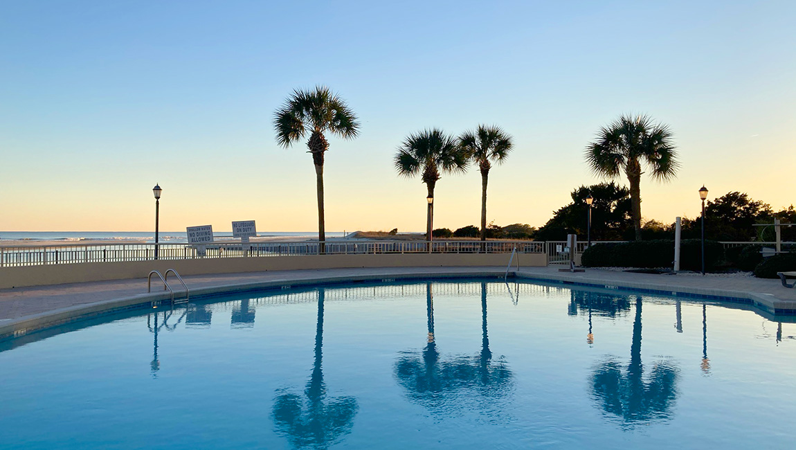 A serene pool area with palm trees and a fence overlooking the beach at sunset.