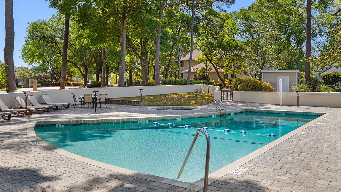 A rectangular pool with a jacuzzi and lounge chairs surrounded by trees and paver stones.