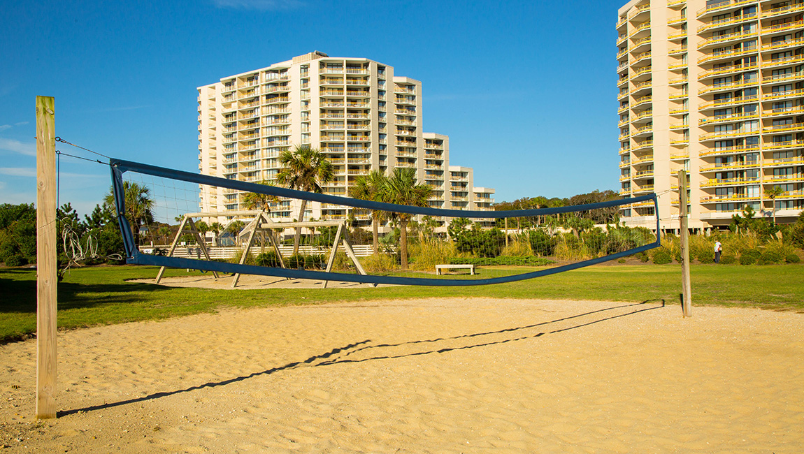 A beach volleyball net on a sandy area with buildings and trees in the background.