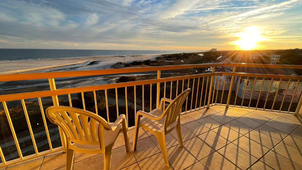 A balcony with a yellow railing and two chairs overlooking the beach at sunset.