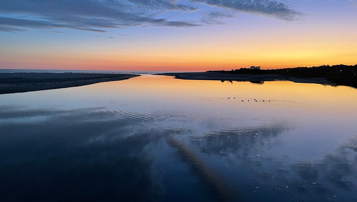 A serene water scene at sunset with birds flying over the water and a reflection of the colorful sky.