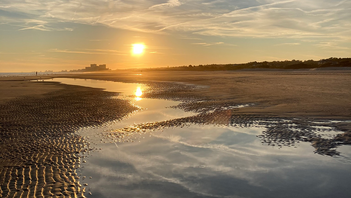 A serene beach scene at sunset with a reflection of the sun and clouds in a puddle of water on the sand.