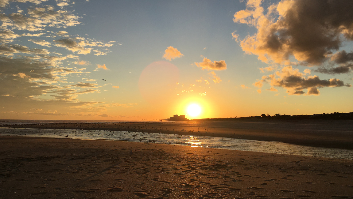 A serene beach scene at sunset with a vibrant orange sky, clouds, and a calm ocean.