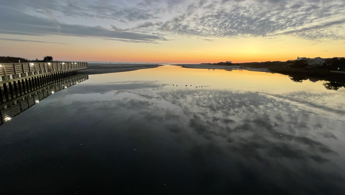 A serene beach access pier at sunset with a calm water reflection.